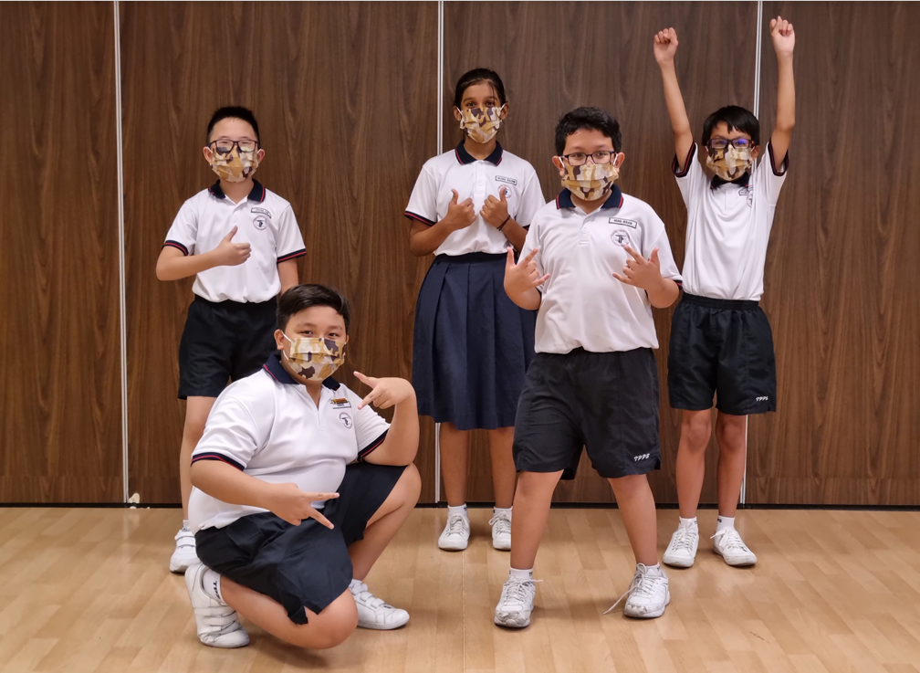 Five children in school uniforms and patterned face masks pose energetically indoors on a wooden floor.